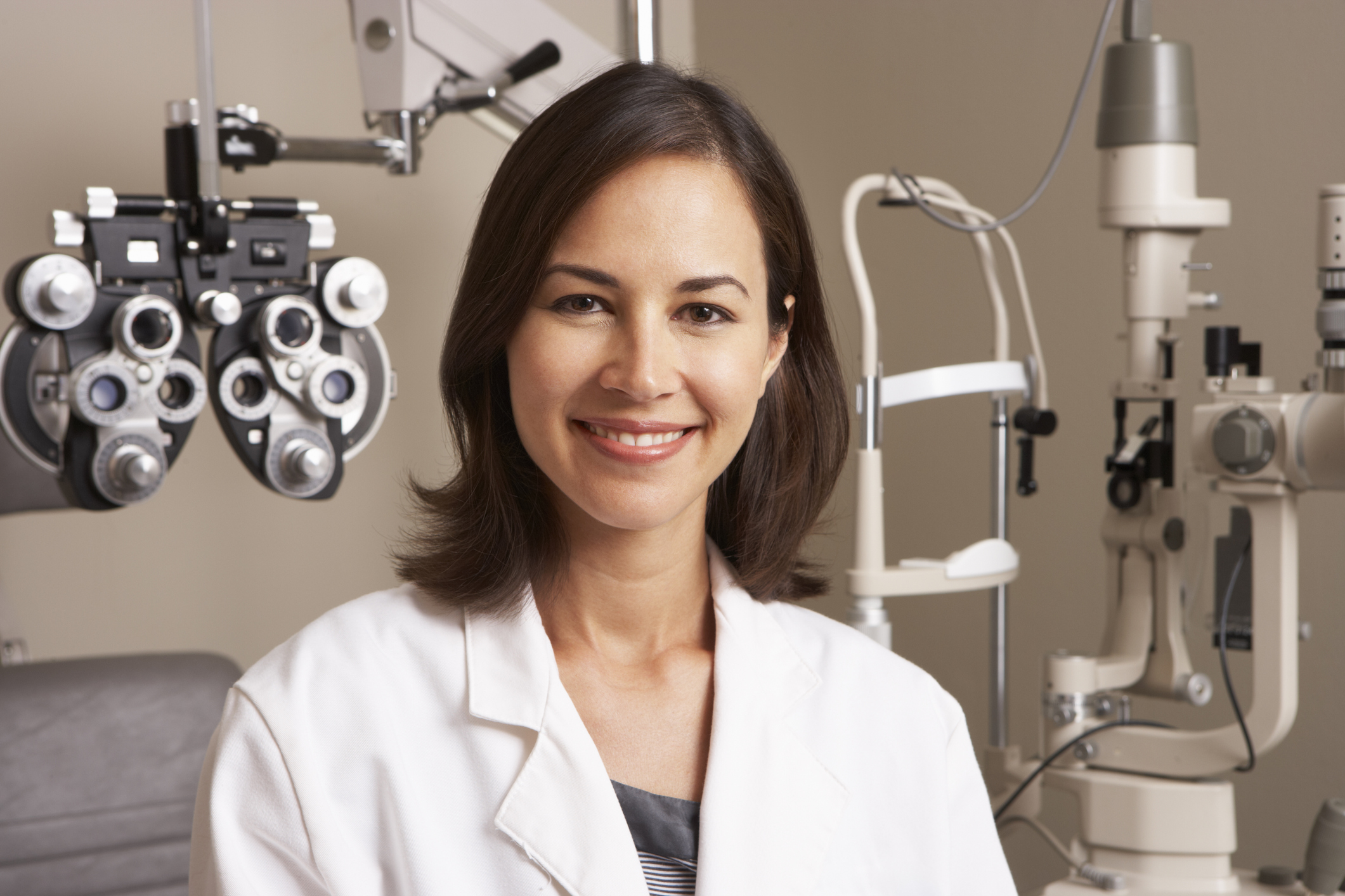 A young female optician in surgery smiling. Varying types of optical equipment fill the background behind the woman eye doctor. She has brunette hair and is wearing a white doctor's coat. The woman is in sharp focus while the background behind her is intentionally blurred.